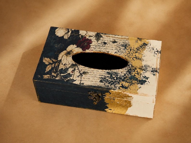 May include: A rectangular wooden tissue box cover featuring a dark blue base with a floral pattern of white and dark red flowers on the left side. The right side displays a distressed white finish with gold leaf accents and a metal latch.