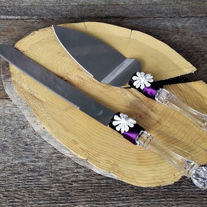 May include: A set of two cake cutting knives with clear glass handles and purple ribbon accents. Each handle is decorated with a white flower.
