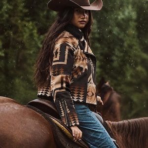 May include: A brown felt cowboy hat and a cropped jacket featuring a geometric pattern in shades of brown, tan, and black. The jacket has a black collar, long sleeves with zippered cuffs, and is paired with blue denim jeans while riding a horse.