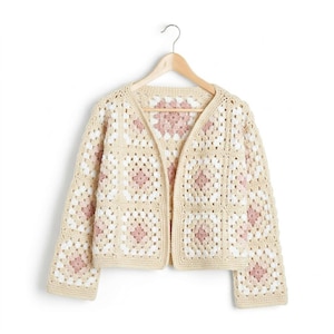 Peut inclure: Un cardigan à manches longues composé de carrés de crochet dans des tons crème, blanc et rose poudré. Le vêtement présente une encolure en V, une ouverture sur le devant et est présenté sur un cintre en bois sur un fond blanc.