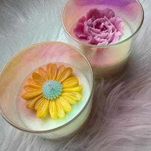 Peut inclure: Deux bougies en verre avec des finitions irisées. L'une présente une décoration en cire en forme de marguerite jaune avec un centre bleu clair. L'autre contient une décoration en cire en forme de pivoine rose. Les deux ont une base en cire blanche.