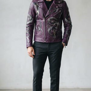 Può includere: Una giacca da motociclista in pelle viola scuro con intricati ricami a serpente argento e nero sul petto e sulle maniche, abbinata a una t-shirt nera, pantaloni grigio scuro e stivali in pelle nera.