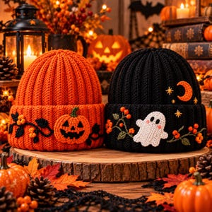 May include: Two cuffed knit beanies, one bright orange and one black. The orange beanie features an embroidered jack-o'-lantern and vine motif. The black beanie features an embroidered white ghost, orange crescent moon, and stars with vine accents.