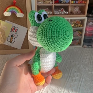Puede incluir: Un peluche tejido a mano de un personaje de dinosaurio verde con un hocico grande y redondeado, vientre blanco y pies naranjas. La figura presenta ojos ovalados negros y blancos, una cabeza verde y un detalle de silla de montar roja en la espalda.
