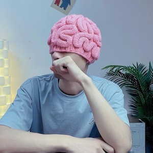 Puede incluir: Un gorro de punto rosa claro con un diseño texturizado en relieve que imita la apariencia de un cerebro humano. El gorro está confeccionado con hilo grueso, creando una serie de crestas sinuosas e interconectadas en toda la superficie.