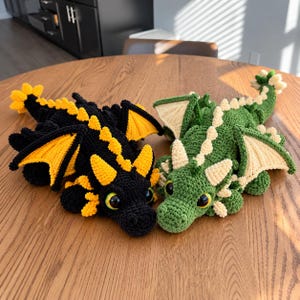 May include: Two handmade crochet dragon plush toys. One dragon is black with bright yellow accents on its wings, horns, and back spikes. The other dragon is green with cream-colored wings, horns, and back spikes. Both have large, glossy eyes.