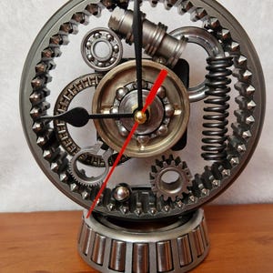 May include: A circular desk clock constructed from various metal mechanical components including gears, ball bearings, and a coiled spring. The clock face features black hour and minute hands and a red second hand, all mounted on a heavy metal base.