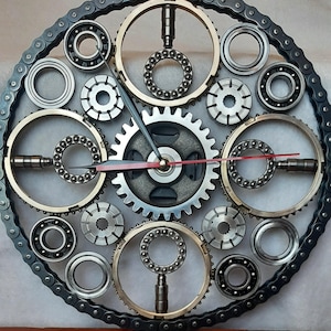 Könnte beinhalten: Eine kreisförmige Wanduhr aus industriellen mechanischen Bauteilen mit einem äußeren Rand aus einer dunklen Fahrradkette, verschiedenen Stahlkugellagern und Zahnrädern. Das Zifferblatt hat zwei dunkelgraue Zeiger und einen roten Sekundenzeiger.