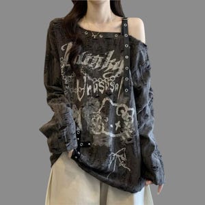 Può includere: Una maglia a maniche lunghe grigio antracite con una texture in tessuto consumato e strappato. Presenta una stampa grafica bianca di un gatto dei cartoni animati, motivi a catena e il testo "Lucky Unusual". Un cinturino nero con fibbia è fissato allo scollo.