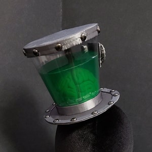 May include: A silver top hat with a clear glass dome containing a green brain. The hat is on a black velvet mannequin head.