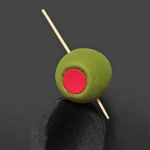 May include: A whimsical olive-themed headpiece. A large, green olive with a red center is impaled by a wooden skewer, perched atop a black mannequin head. The olive is the focal point, set against a dark background.