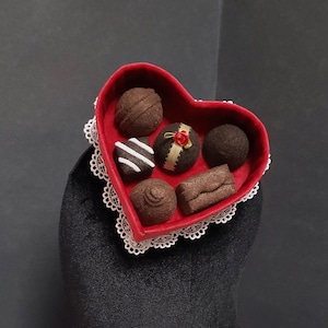 Könnte beinhalten: Eine rote Filzbox in Herzform mit einer weißen Spitzenborte, gefüllt mit Schokoladenbonbons. Die Box sitzt auf einem schwarzen Samt-Mannequin-Kopf.