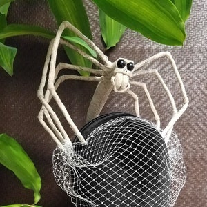 May include: A white spider hat with a black veil. The spider has large, black eyes and a white body. The hat is sitting on a black mannequin head.