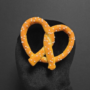 May include: A pretzel shaped hair clip with a brown and yellow color scheme. The pretzel is covered in small white sugar crystals.