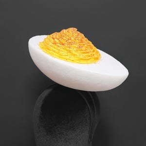 Op de afbeelding: Een grillige hoed in de vorm van een gevuld ei, met een witte eierschaal en een gele dooier met rode stippen. De hoed staat op een zwarte mannequinhoofd tegen een donkere achtergrond.