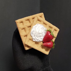 May include: A brown waffle hat with white frosting and red strawberries on top. The hat is on a black mannequin head.