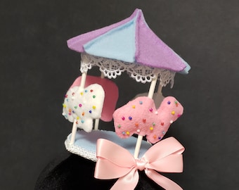 May include: A whimsical carousel-themed hair accessory. The miniature carousel features a light blue and purple roof, white lace trim, and pink and white felt animal figures decorated with colorful sprinkles. A pink satin bow is attached at the base.