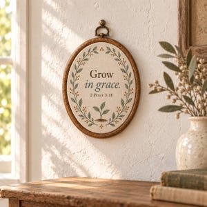 May include: An oval wall hanging featuring a wooden frame and a fabric centre. The fabric displays a green leaf wreath, a small sprout illustration, and the text "Grow in grace. 2 Peter 3:18" in dark brown lettering. A metal hanger is at the top.