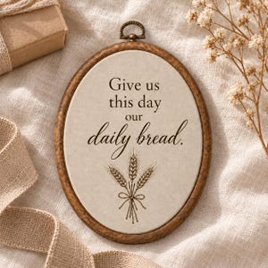 Peut inclure: Une décoration murale ovale dotée d'un cadre couleur bois brun foncé avec une boucle en métal sur le dessus. Le panneau central est blanc cassé avec le texte "Give us this day our daily bread." et une illustration détaillée de blé.