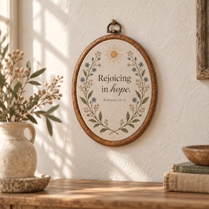 May include: An oval wall hanging featuring a wooden frame and a fabric centre with the text "Rejoicing in hope. Romans 12:12". The design includes a sun illustration at the top and floral sprigs with blue and yellow flowers along the sides.
