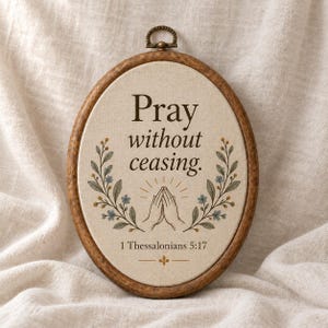 May include: An oval embroidery hoop frame containing a cream-colored fabric panel. The text reads Pray without ceasing. 1 Thessalonians 5:17. Below the text is an illustration of two hands pressed together in prayer, flanked by floral sprigs.