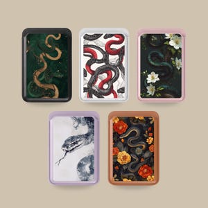 May include: Five rectangular magnetic card wallets featuring distinct snake-themed designs. The collection includes a black, white, pink, lavender, and brown wallet, each displaying intricate illustrations of snakes, flowers, and abstract patterns.