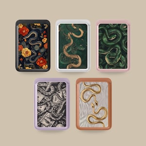 Può includere: Cinque porta carte magnetici rettangolari con angoli arrotondati. La fila superiore include un porta carte nero con serpenti e fiori, uno bianco con un serpente verde e uno rosa con serpenti verdi. La fila inferiore presenta un porta carte color lavanda con serpenti e banconote, e uno color cuoio con due serpenti dorati.