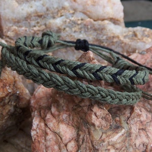 Könnte beinhalten: Zwei geflochtene Armbänder, eines grün und eines schwarz mit grünen Streifen. Die Armbänder sind miteinander verbunden und liegen auf einem Stein.