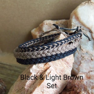 May include: Two braided bracelets, one black and one light brown, are displayed on a rock. The bracelets are made of woven cord and are a simple, stylish accessory. Black & Light Brown Set.