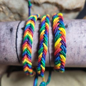 May include: Three braided friendship bracelets in a rainbow of colors: red, orange, yellow, green, blue, and purple. The bracelets are made of woven threads and are displayed on a neutral-toned surface. The bracelets are a vibrant accessory.