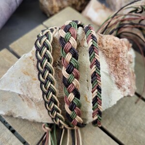 May include: Three braided bracelets in brown, green, and beige colors. The bracelets are made of woven cord and have tassels at the ends.