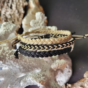 Könnte beinhalten: Drei geflochtene Armbänder, eines schwarz und zwei beige, liegen übereinander. Die Armbänder sind aus geflochtener Schnur gefertigt.