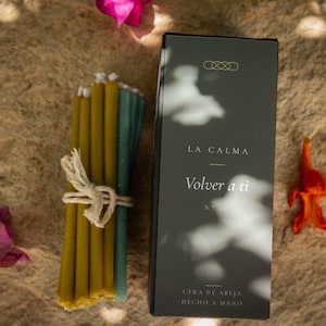 May include: A bundle of thin, mustard yellow and teal beeswax candles tied with a white string, positioned next to a rectangular dark green box with the text LA CALMA, Volver a ti, N° 01, CERA DE ABEJA, HECHO A MANO printed in white.