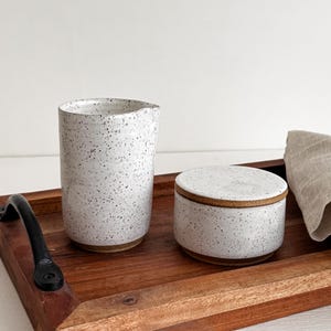 Puede incluir: Una bandeja de madera con un asa negra sostiene una taza de cerámica blanca moteada y un pequeño recipiente redondo con tapa. Una servilleta de lino doblada descansa sobre la bandeja. La bandeja es de madera oscura.