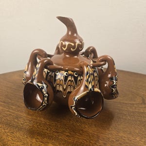 May include: A brown ceramic vessel with a central lidded container and six attached small cups. The piece features a marbled, wavy pattern in cream, dark blue, and black around the center, with a smooth brown finish on the lid and handles.