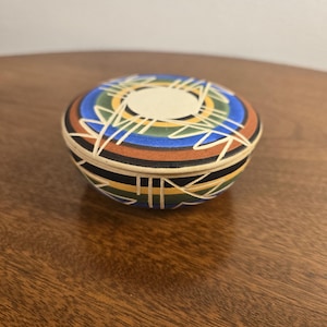 May include: A small, round, lidded ceramic bowl featuring a matte finish with concentric bands of blue, green, black, and terracotta. The surface is decorated with white, incised geometric line patterns that radiate from a central cream circle.