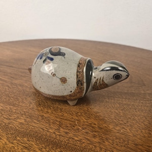 Pode incluir: Uma pequena estatueta de tartaruga em cerâmica com corpo cinza-claro salpicado. O casco apresenta motivos florais pintados à mão em azul e castanho. Uma faixa castanha escura separa o casco da cabeça, que tem detalhes em preto e castanho.