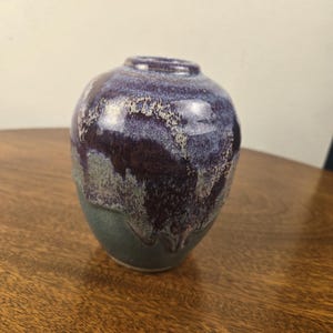 May include: A small, rounded ceramic vase with a narrow opening. The upper portion features a glossy, deep purple glaze that drips down over a matte, dark gray base, creating a textured, layered appearance with visible color blending.