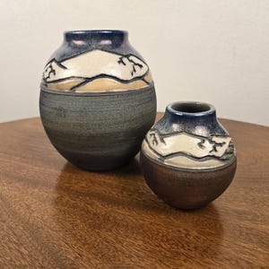 May include: Two round ceramic vases with narrow openings. The larger vase has a dark grey base and a blue rim with a white mountain range design. The smaller vase features a dark brown base and a similar blue rim with a white mountain range pattern.