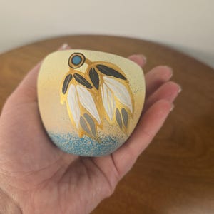 Pode incluir: Uma pequena tigela de cerâmica arredondada com uma base de cor creme que se desvanece para um fundo azul salpicado. O exterior apresenta um design pintado à mão de duas penas brancas e cinzentas delineadas a ouro, com um pequeno círculo turquesa.