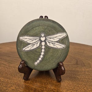 May include: A circular, dark olive green ceramic tile featuring a white, embossed dragonfly design with a heart-shaped thorax. The tile is displayed on a small, dark brown wooden easel stand, showcasing a textured surface and detailed insect wings.