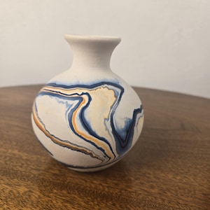 May include: A small, round ceramic vase with a matte white finish, featuring swirling marbled patterns in shades of deep blue, soft orange, and grey. The vase has a narrow neck and a flared rim, with a smooth, rounded body shape.