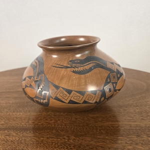 May include: A rounded terracotta-colored clay pot featuring a wide opening and a tapered base. The exterior surface is decorated with a black painted design depicting a stylized snake with geometric patterns integrated into its elongated body.