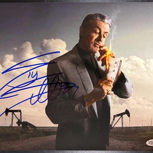 May include: A glossy photograph featuring Sylvester Stallone in a grey suit and black shirt, lighting a document on fire. The image includes a bold blue autograph across the left side and a Total Sports Authentics sticker in the corner.