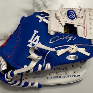 Puede incluir: Un guante de béisbol Rawlings azul y blanco con el logotipo LA de los Dodgers de Los Ángeles, una firma plateada y una pegatina holográfica de Total Sports Authentics. El guante tiene la etiqueta 10 Inch y THE GOLD GLOVE CO. TM con costuras rojas.