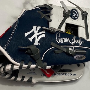 Puede incluir: Un guante de béisbol azul marino y blanco con el logotipo de los New York Yankees en blanco, una firma plateada y el logotipo de Rawlings en la muñequera. El guante incluye cordones grises, una etiqueta roja y una pegatina de autenticación.