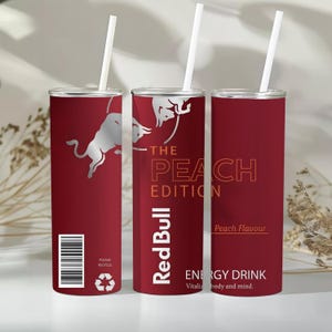 Könnte beinhalten: Drei zylindrische Becher in Dunkelrot mit weißen Strohhalmen. Das Design zeigt das silberne Red Bull Logo, den Text THE PEACH EDITION, Red Bull, Peach Flavour, ENERGY DRINK, Vitalizes body and mind, einen Strichcode und ein Recyclingsymbol.
