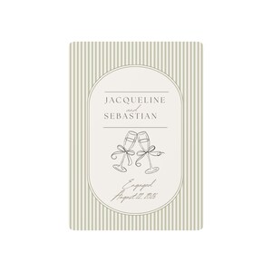 May include: A rectangular engagement announcement card featuring a beige and white vertical striped border. The centre contains an oval frame with the text JACQUELINE and SEBASTIAN, a line drawing of two champagne flutes with bows, and Engaged August 22, 2026.