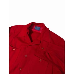 May include: A bright red long-sleeve shirt featuring a pointed collar, a front button-down closure, and two chest pockets with button flaps. A blue Pendleton brand label is stitched onto the inside of the collar area of the red garment.