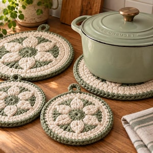 May include: Three round, crocheted pot holders featuring a cream-coloured floral pattern on a sage green background, each with a small hanging loop. A sage green enamelled cast iron pot with a lid sits on one of the pot holders on a wooden surface.